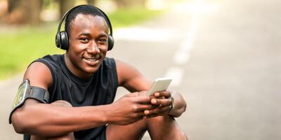 Closeup of smiling black sportsman with wireless headphones using mobile phone while resting on park path, copy space photo