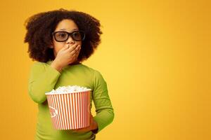 Scary Movie. Shocked little african girl in 3d glasses and with popcorn, covering mouth with hand and looking with frightened face expression photo