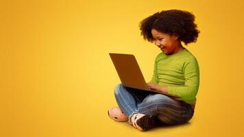Modern Technologies For Kids. Smiling black girl sitting with laptop on lap, playing online games, relaxing on floor over yellow background photo