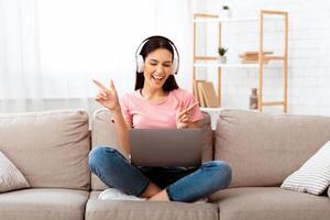 Break From Work Or Study For Favorite Song. Overjoyed emotional young lady listening to music wearing wireless headset, sitting on the sofa at home with computer on her lap and dancing, copy space photo