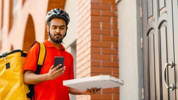 Pizza delivery with man on bike. Serious delivery man with beard in helmet, with yellow backpack holding box and looking at smartphone, near front door, free space photo