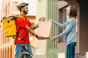 Food, parcels delivery concept. Smiling millennial indian male with beard, smart watch in safety helmet in uniform with yellow thermal bag gives package to woman client near house, side view, panorama photo
