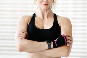 Fitness model and motivation. Friendly adult woman in sportswear with smart watch with arms crossed, demonstrates muscles at home or gym, copy space photo