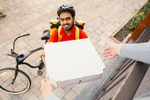 Online order and pizza at home. Smiling courier with beard in helmet gives boxes to client, near house on street, top view, pov photo