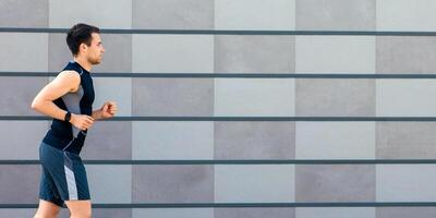 Jogging in city. Slender guy in sportswear with fitness tracker runs on gray brick wall background, side view, panorama, free space photo