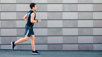 Running in morning in city. Guy in sportswear and with fitness tracker on hand runs at path along stone wall, free space photo