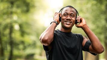 Music for exercising. Joyful black jogger putting wireless headset on his head, training at park, looking at free space photo