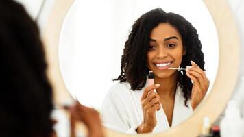 Makeup Routine. Smiling African American Girl Applying Lip Gloss, Looking In Mirror At Bathroom, Getting Ready After Shower, Over Shoulder View photo