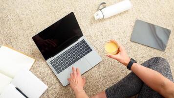 Healthy lifestyle, blog and sport. Woman with smart watch holds glass of orange juice and works on laptop, sitting on floor, with notepads, tablet and bottle, cropped, free space photo