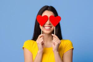 All You Need Is Love. Romantic asian woman posing with red hearts over eyes and smiling, having fun, celebrating valentine's day alone, standing over purple studio background, closeup shot, free space photo