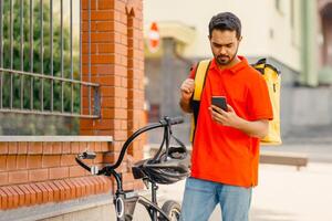 GPS map, coordinates of client's address, delivery service and online order. Concentrated young arabian male courier in orange t-shirt with bicycle and big thermo bag looks in phone, empty space photo