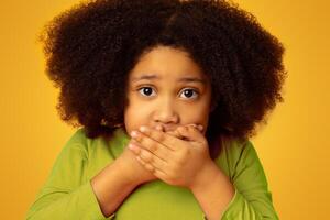Keep Silent. Frightened little girl covering mouth with hands and looking at camera with scared face expression, posing over yellow background photo