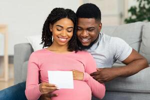 Smiling pregnant black couple sitting by couch and holding white empty card. Cheerful african american husband and wife hugging and looking at empty card, sitting on floor at living room, empty space photo