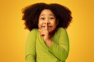 Keep Silence. Funny Little Black Girl Showing Shh Sign, Holding Finger On Mouth And Looking At Camera, Standing Over Yellow Background, Copy Space photo