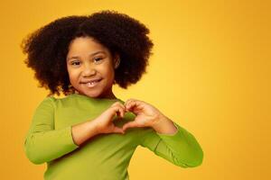 Cute little african american girl making heart shape with her hands and smiling to camera, posing over yellow background in studio, empty space photo