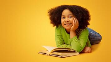 Cute Little African American Girl Reading Book On Floor In Studio And Smiling At Camera, Lying On Yellow Background, Free Space photo