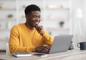 Happy black guy project manager sitting at workdesk at office, typing on laptop keyboard and smiling, chatting with clients, enjoying his job, copy space. Man freelancer working from home photo