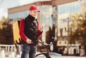 Courier With Phone Using Meal Delivery Application Standing With Bicycle Outside In Urban Area. Business Lunch Delivery. Free Space photo