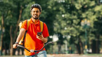 Quick service. Friendly courier at park, in helmet with backpack on bicycle, holding smartphone and looking for client address, free space photo
