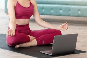 Young lady sitting in lotus pose in front of laptop computer, meditating or doing breath exercises to online tutorial at home photo