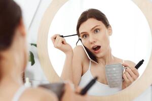 Amazed girl talking on phone with girlfriend while putting mascara in front of mirror at home, drinking morning coffee photo