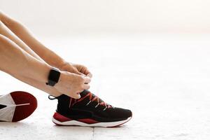Healthy life, keep fit concept. Woman with fitness tracker tying shoelaces on fashion sneakers, sitting on floor in gym or living room, cropped, copy space photo