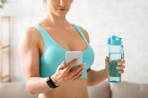 Quality workout online. Adult woman in sport bra with bottle of water and smartphone in hand, looks at result in phone, copy space photo