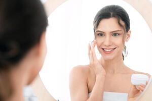 Good looking young lady using face cream at home, looking at mirror and smiling, shot from behind. Smiling woman with ponytail holding cream jar and applying beauty product on her cheeks photo
