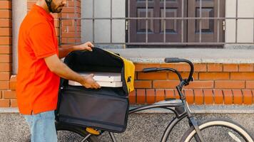 Delivery home and office. Young delivery man in helmet takes out pizza from big yellow backpack on bicycle, near front door, side view, free space photo