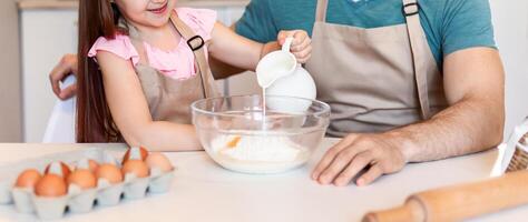 Cheerful Dad And Daughter Baking Cake, Little Girl Adding Milk To Dough Cooking With Daddy In Kitchen At Home. Family Of Two Enjoying Pastry Preparation On Weekend. Selective Focus photo