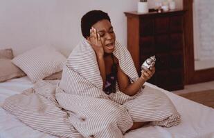 Suffering from pain, waking up in morning, back to work, tired after sleep. Sad millennial african american lady presses her hand to temple and sits on bed in blanket, holds alarm clock and screams photo