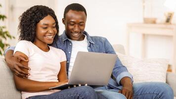 Smiling black couple using laptop while resting on couch at living room at home, watching funny content together, copy space photo