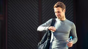 Athletic style and preparation for run. Young smiling man goes to training with sports bag and in sportswear, looks at tracker on hand and holds water photo