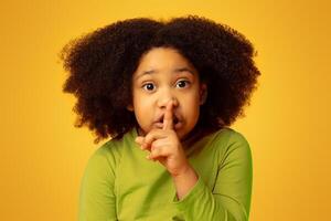 Shh. Serious little african girl showing silence sign, asking be quiet, isolated on yellow background with free space photo