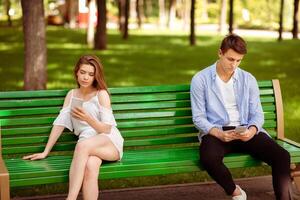 Gadget addiction and relationship problems. Millennial couple with smartphones sitting on bench at park, ignoring each other photo