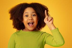 I Have Idea. Little african american girl pointing finger up and looking at camera, standing over yellow studio background with free space photo