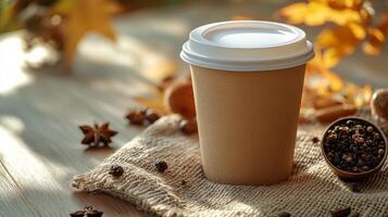 A brown disposable cup sits on a burlap cloth, surrounded by colorful autumn leaves and an assortment of spices on a wooden table, evoking a warm, cozy atmosphere. photo