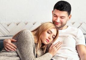 A young couple enjoys a quiet moment together on a sofa, sharing affection and warmth. The woman rests her head on the man's shoulder, creating a peaceful, intimate atmosphere. photo
