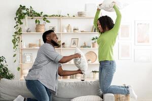 A joyful black couple engages in pillow fight, laughing and playfully challenging each other. The cozy living room is filled with plants and decorative items, enhancing the cheerful atmosphere. photo