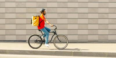 Deliveryman on bicycle. Young guy with beard and big backpack rides along path in city on gray brick wall background, side view, free space photo