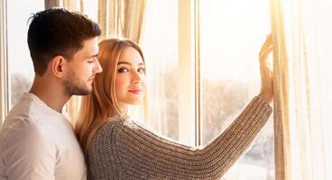 A couple stands together by bright, large windows, adjusting light curtains. The warm sunlight creates a romantic atmosphere, enhancing their intimate moment and cozy setting. photo