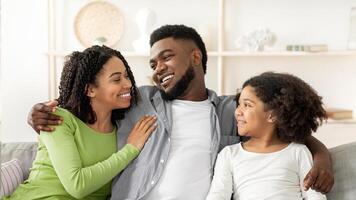 A joyful black family sits closely on a couch in their living room, sharing smiles and laughter. The warm afternoon light enhances their bond and creates a delightful atmosphere. photo