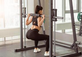 A woman in athletic wear demonstrates proper squat technique using a barbell in a gym setting. The well-lit space emphasizes a focus on strength training in the morning. photo