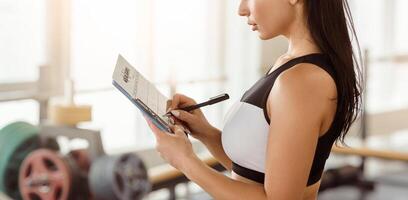 A fitness enthusiast stands in a gym, taking notes on her workout routine. The setting features exercise equipment and weights, creating a focused environment for training. photo