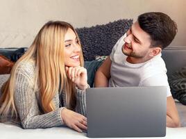 A young couple lies comfortably on a couch, smiling and looking at a laptop together. They share a cozy moment, engaging with each other and the content on the screen. photo