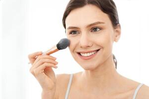 Closeup of attractive young woman applying blush on her face, sitting in front of mirror, copy space. Smiling attractive young lady doing makeup before going out, shot over shoulder in bedroom photo