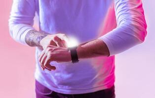 A man with a tattoo adjusts his smartwatch while standing against a vibrant, colorful backdrop. It captures a modern moment of technology integration in daily life during the evening. photo