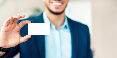 A young Middle Eastern businessman stands in an office, smiling confidently while holding up a blank business card. The setting reflects a corporate atmosphere, ideal for networking. photo