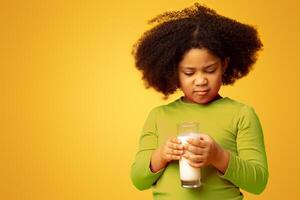 Grumpy little girl refusing to drink milk, looking at glass with disgust, standing over yellow studio background with free space photo