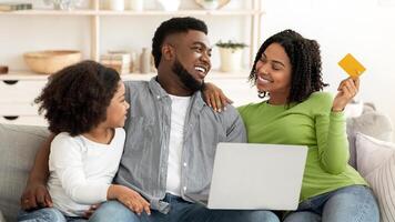 A black family is gathered in their cozy living room, happily engaging in online shopping while sharing smiles and laughter. The child sits beside the parents, creating a warm atmosphere. photo